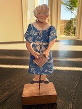 A handcrafted wooden figurine of an elderly woman with gray hair and glasses, wearing a blue and white patterned dress, standing on a wooden base.