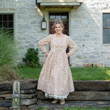Melissa wearing a long-sleeved, knee-length floral dress stands outdoors in front of a stone house, with tall ornamental grasses and a rustic stone wall nearby.