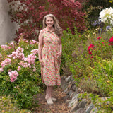 Melissa stands on a stone path in a lush garden, wearing a sleeveless, knee-length floral dress with a red, pink, and beige pattern. The background features a variety of colorful flowers, green foliage, and trees, with house walls visible on both sides.