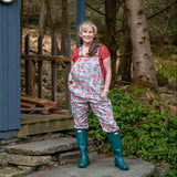 Melissa stands on the stone steps of a blue cabin wearing floral print overalls, a red t-shirt, and green rubber boots, with trees and greenery in the background.