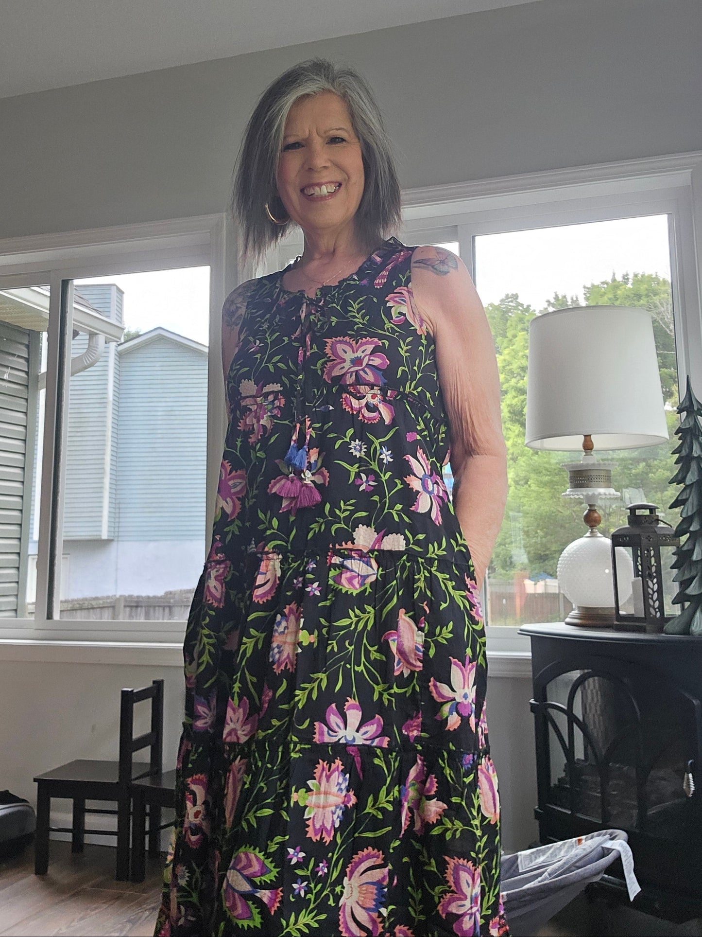 A woman wearing a sleeveless black dress with a vibrant pink, purple, and green floral pattern, standing in a home interior near a window.