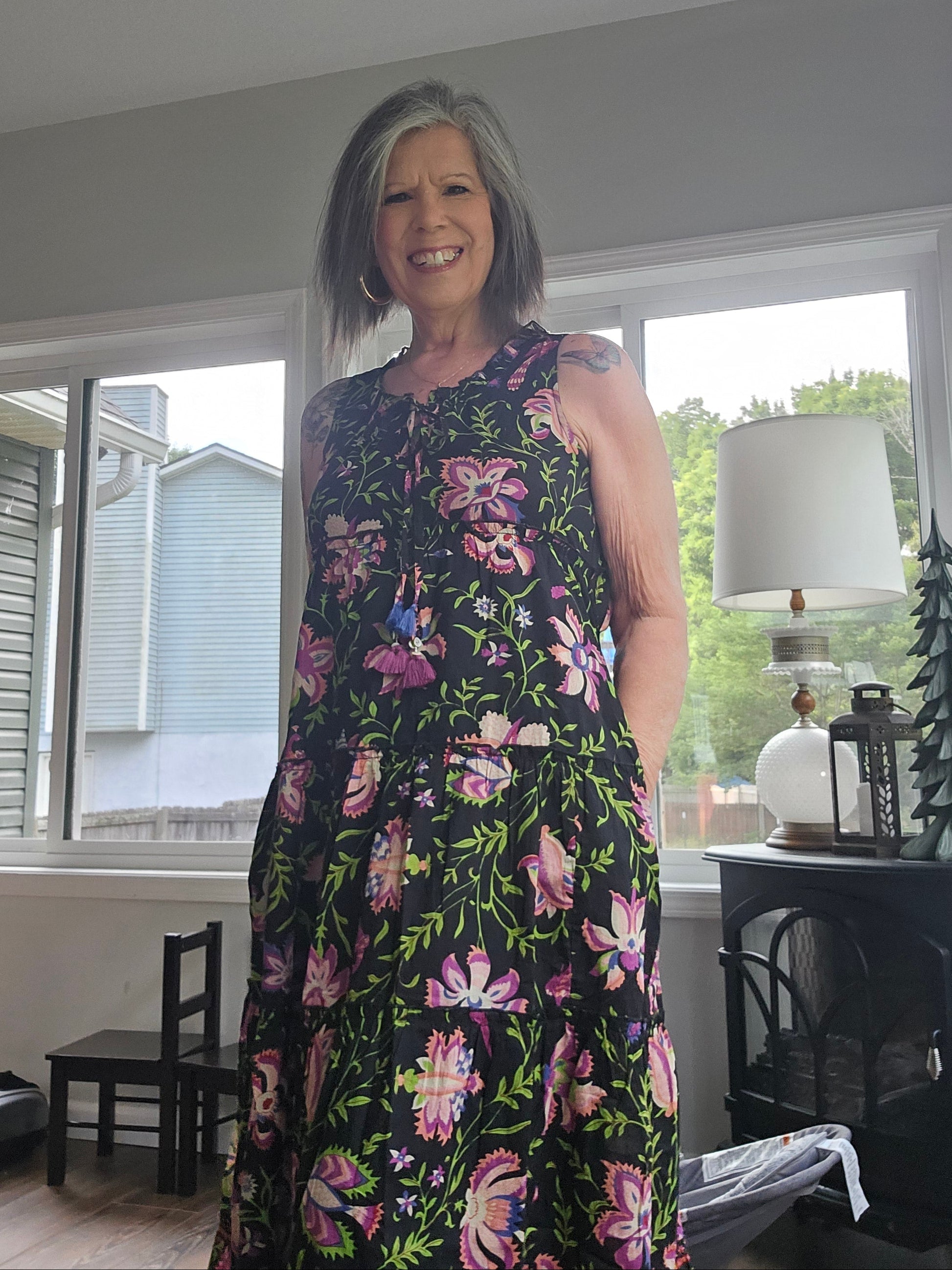 A woman wearing a sleeveless black dress with a vibrant pink, purple, and green floral pattern, standing in a home interior near a window.