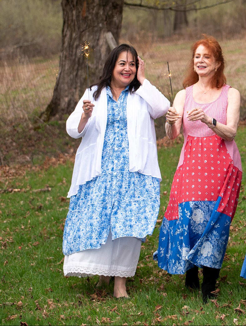 Two women walking outdoors in a wooded park setting, each wearing a different colorful dress and cardigan outfit. The outfits include red, blue, and white tones, and the background features grass, trees, and a rustic fence.