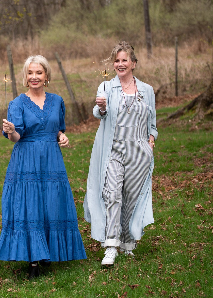 Two women walking outdoors in a wooded park setting, each wearing a different colorful dress and cardigan outfit. The outfits include blue, and gray tones, and the background features grass, trees, and a rustic fence.