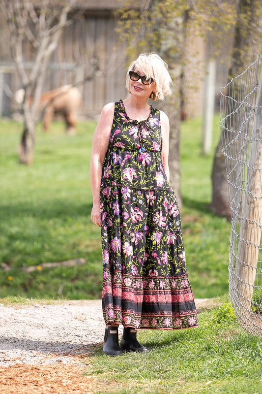 A woman stands on a dirt path at a farm wearing a sleeveless black maxi dress with a pink and green floral pattern and black boots. Trees, a wire fence, and a grassy field are visible in the background.