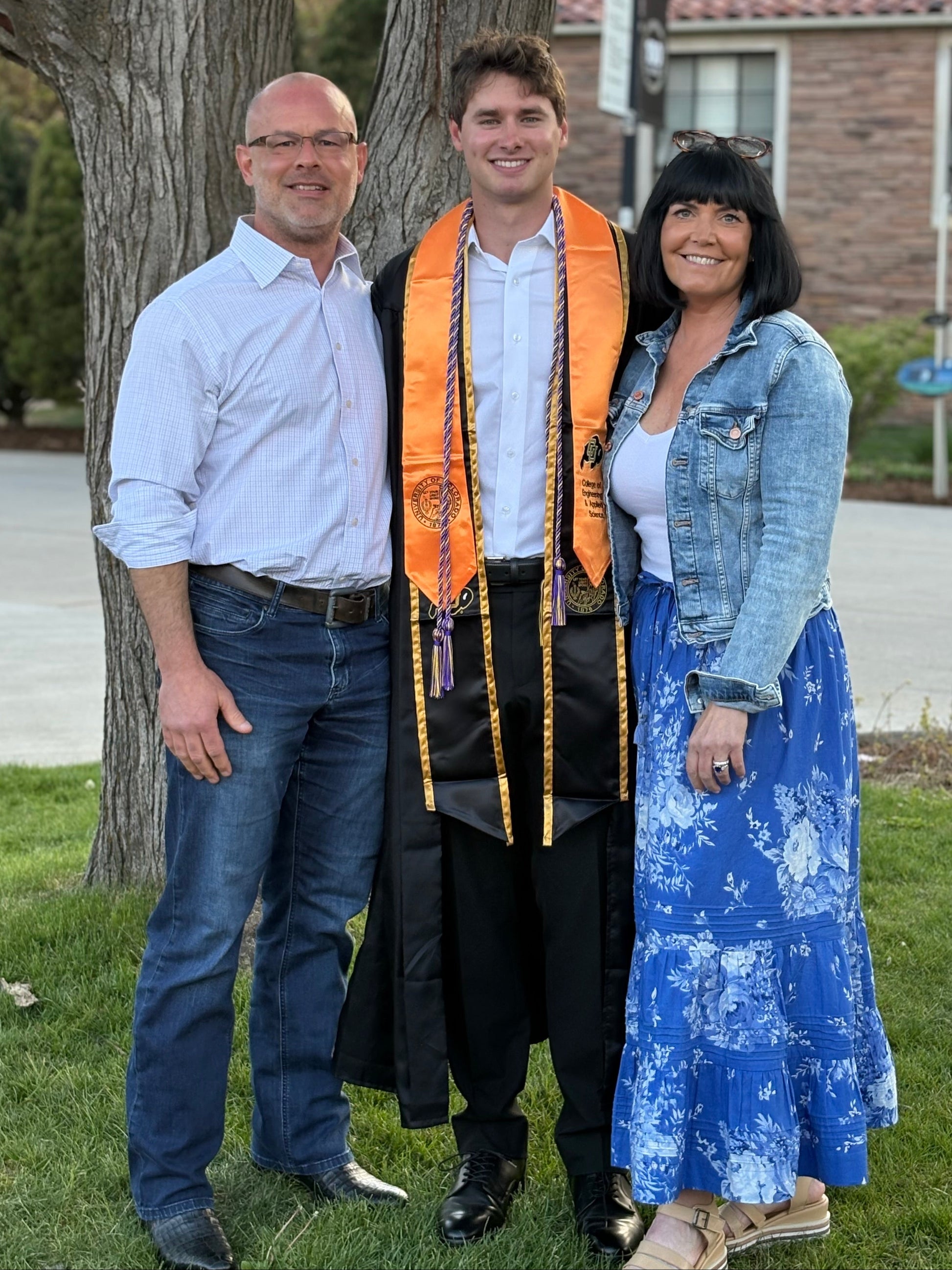 Three people standing outdoors in front of a tree and brick house. The central person is wearing a graduation gown and an orange and gold sash, flanked by a man in a light blue shirt and jeans, and Nicole in a denim jacket and blue floral skirt.