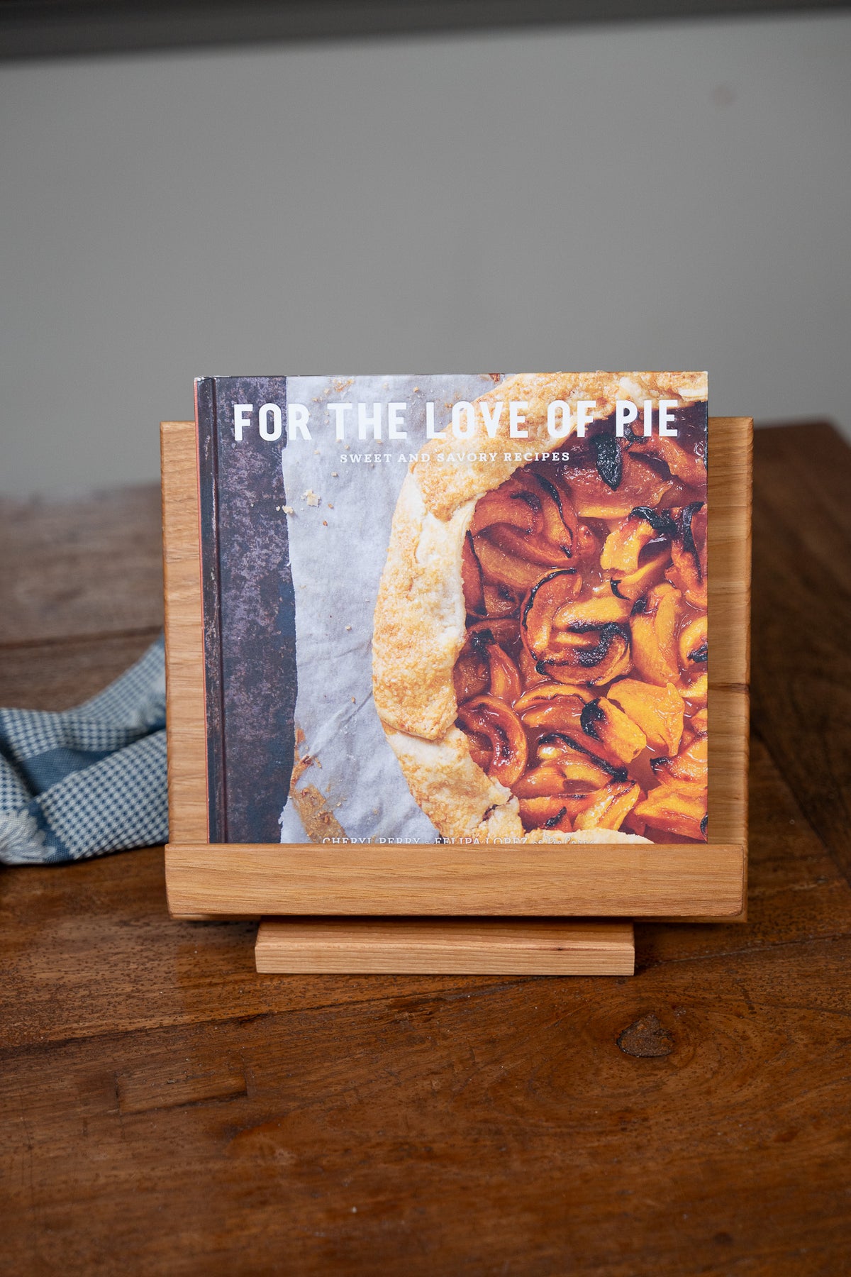 A cookbook titled 'FOR THE LOVE OF PIE' displayed upright on a small wooden stand on a wooden table, with a blurred kitchen background.