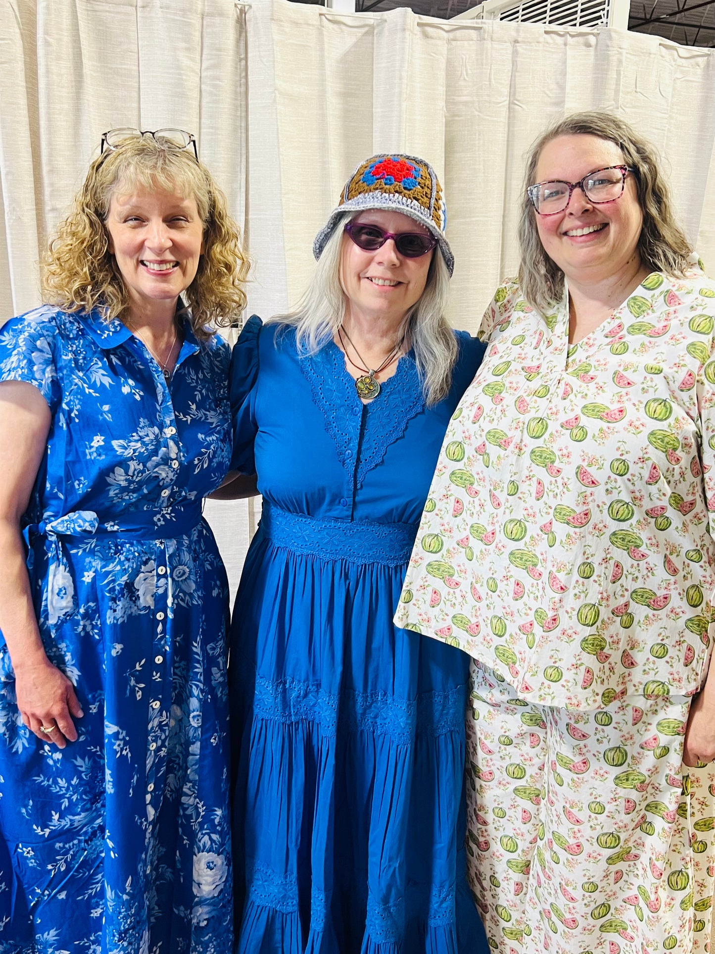 Three women standing together and smiling, wearing colorful patterned dresses. The woman on the left wears a royal blue floral dress, the woman in the center wears a bright blue tiered dress with a patterned hat, and the woman on the right wears a cream dress with a green and pink paisley pattern.
