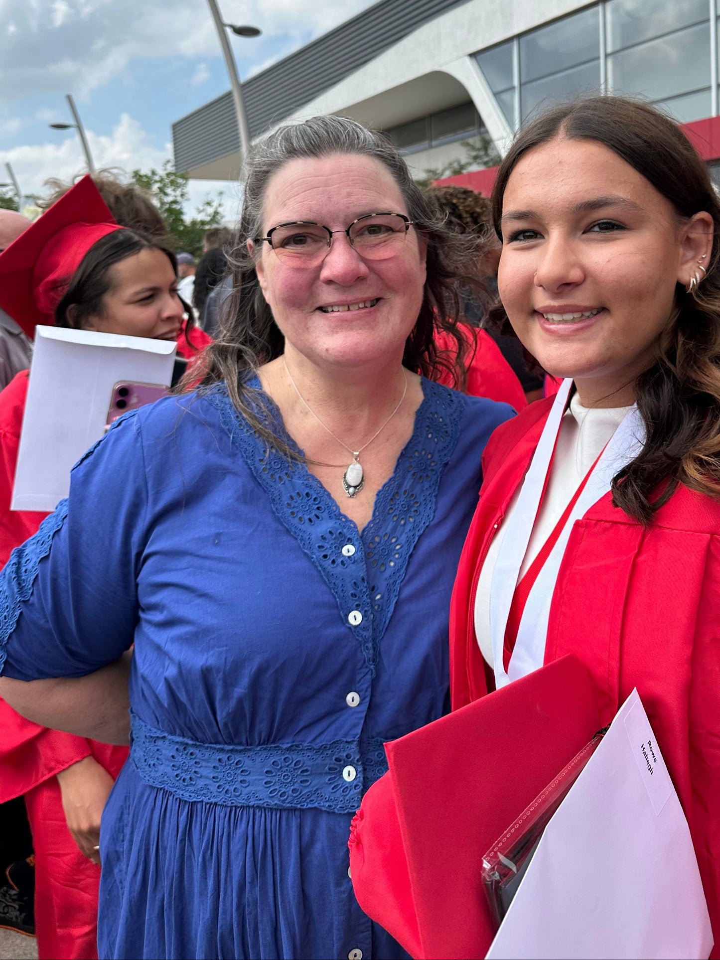A group of graduates wearing red and blue graduation gowns and caps, smiling outdoors in front of a modern building with a visible 'WAMBO' sign.