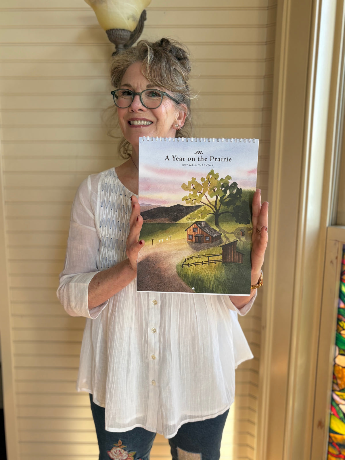 Melissa holds up a spiral-bound book titled 'A Year on the Prairie' by Jane Cochrane. The book cover features a colorful illustration of a rural prairie landscape with a cabin, tree, and fields.