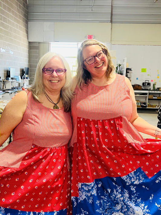 Two women smiling and posing indoors while wearing matching sleeveless dresses with a red gingham bodice, red patterned midsection, and blue floral lower skirt.