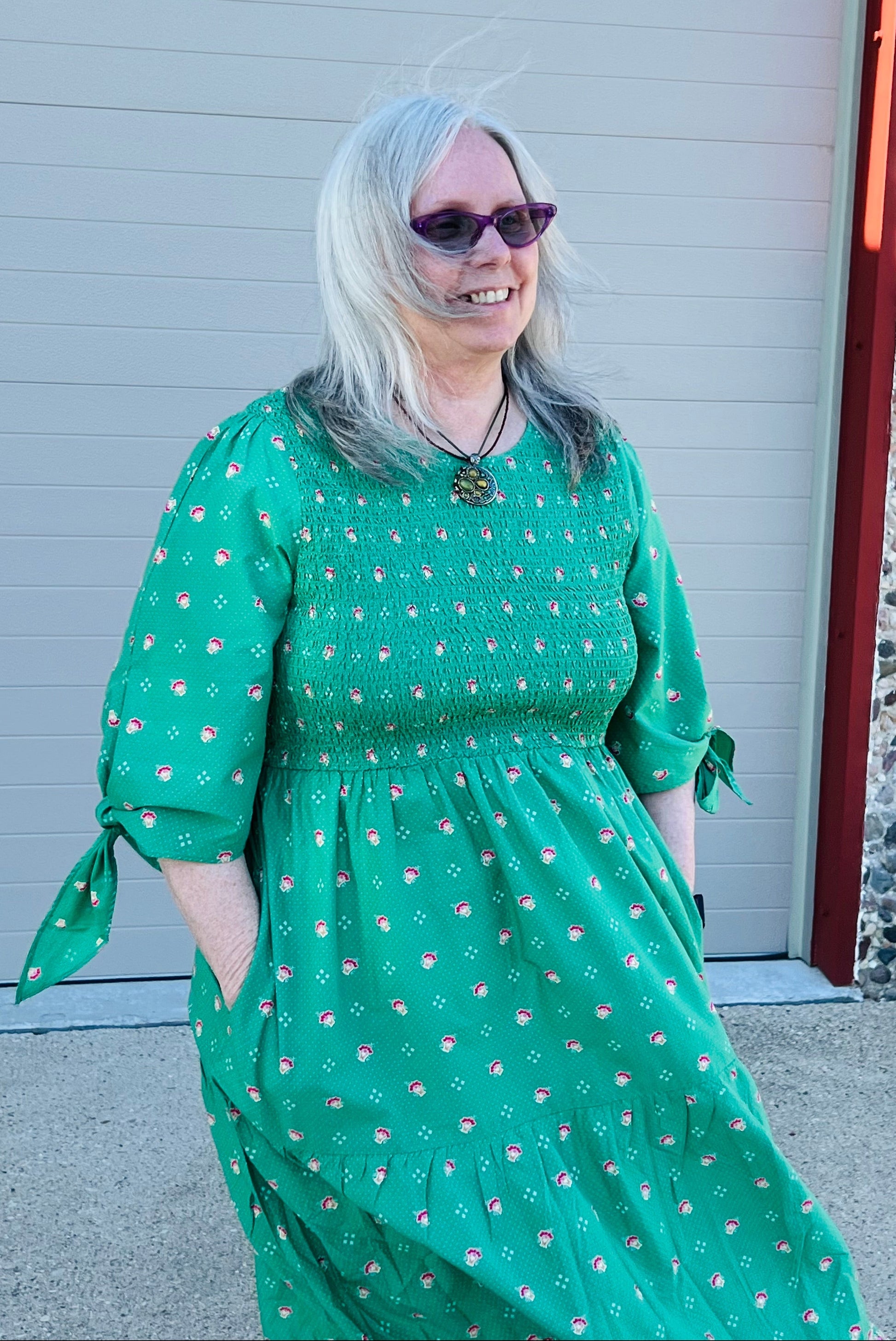 A woman with gray hair and sunglasses wearing a green tiered midi dress with a small pink and white floral pattern, standing in front of a gray garage door.