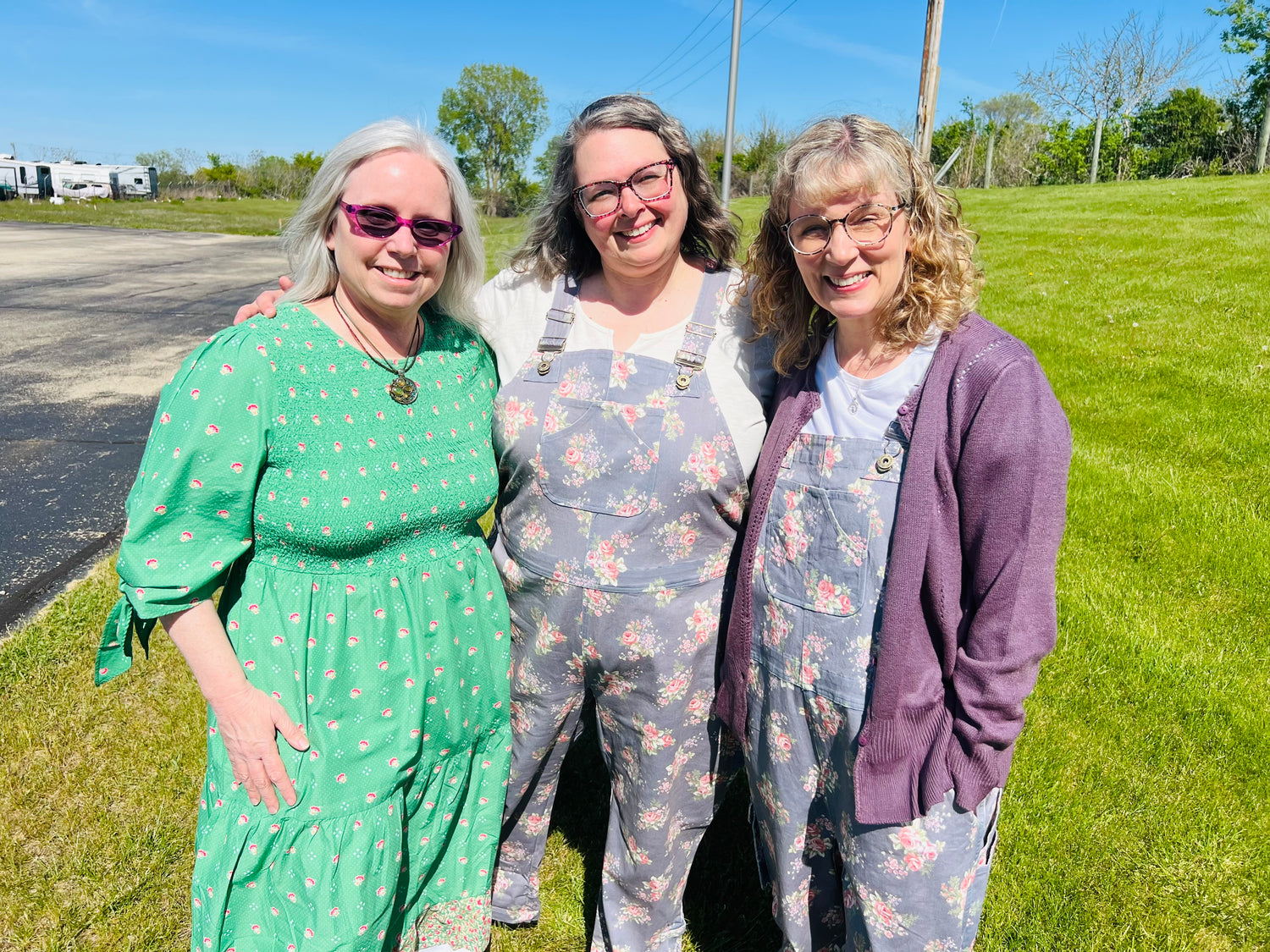 Three women standing together outdoors on a grassy field under a blue sky. The woman on the left wears a green dress with white polka dots and pink bow details, while the other two wear matching floral overalls.