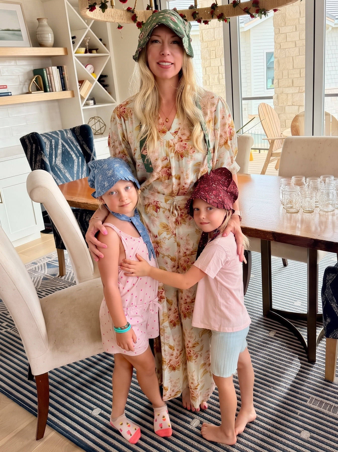 A woman wearing floral pajamas and green bonnet stands with two young girls in a bright dining room. The girls are wearing pastel outfits and bonnets.