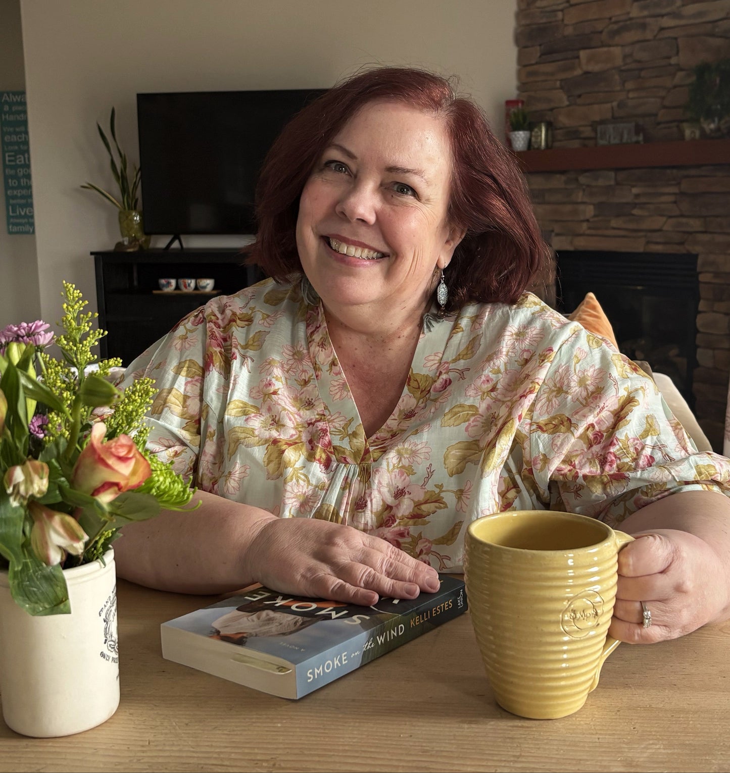 A smiling woman with red hair sits at a wooden table in a cozy living room setting. She is wearing a light floral print blouse and has a yellow ribbed ceramic mug in front of her, with a book and a vase of flowers on the table.