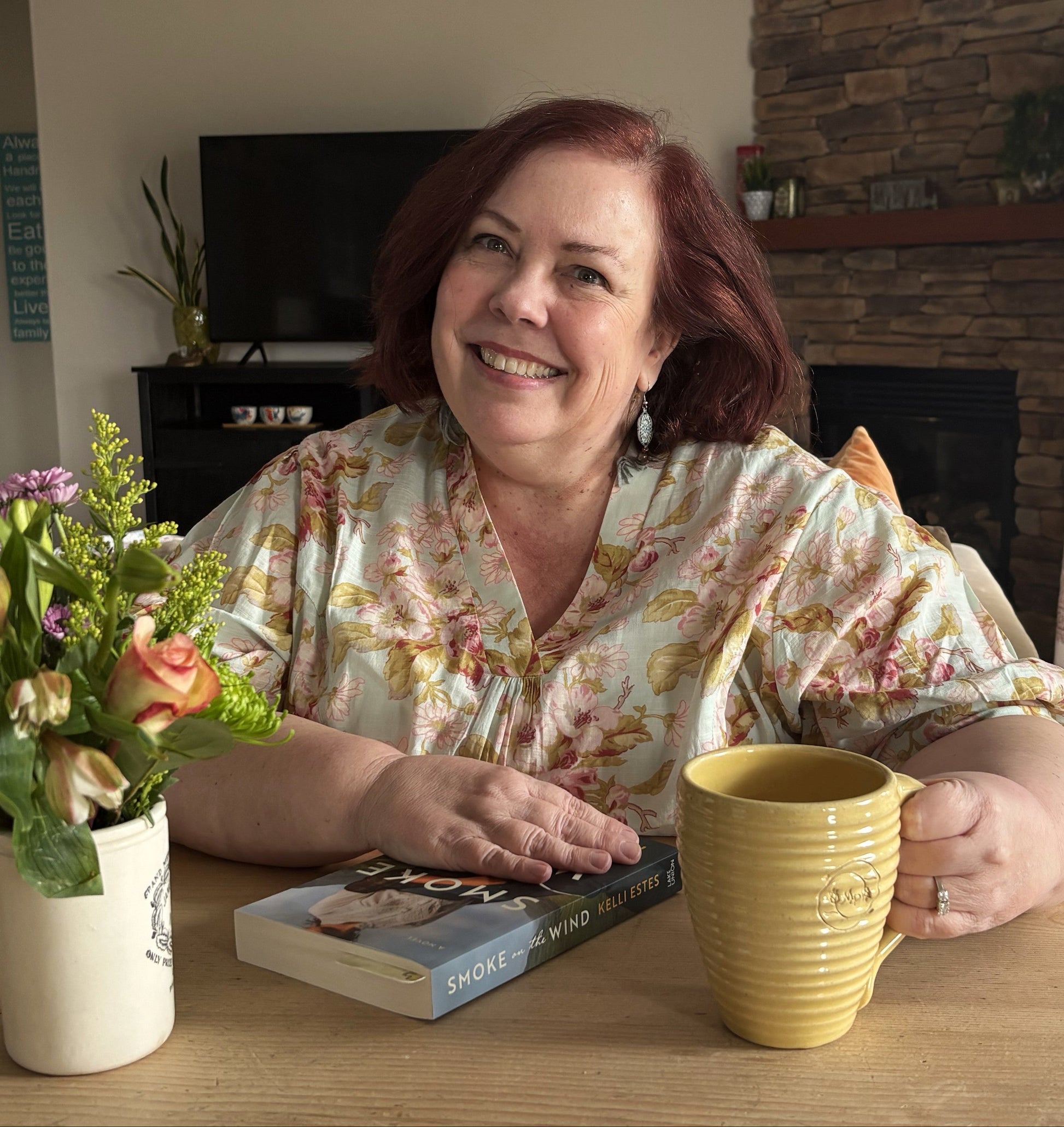 A smiling woman with red hair sits at a wooden table in a cozy living room setting. She is wearing a light floral print blouse and has a yellow ribbed ceramic mug in front of her, with a book and a vase of flowers on the table.