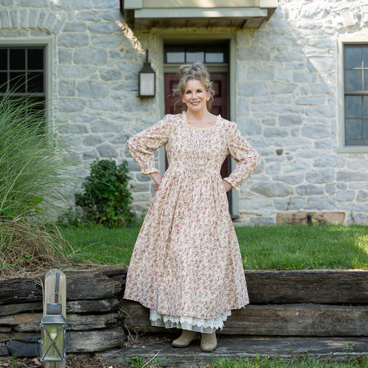 Melissa wearing a long-sleeved, knee-length floral dress stands outdoors in front of a stone house, with tall ornamental grasses and a rustic stone wall nearby.