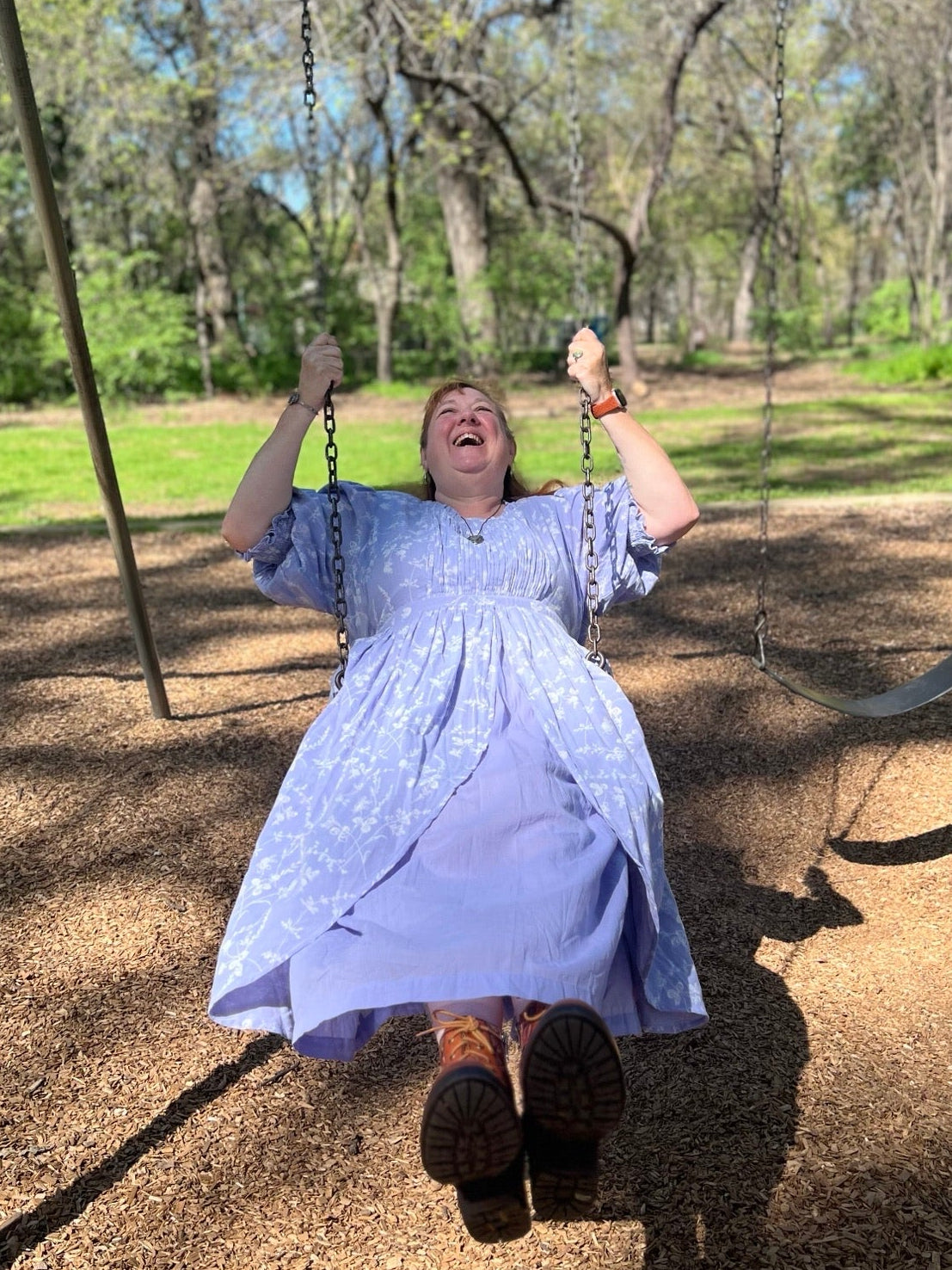 A person wearing a light lavender-blue patterned dress swings on a playground swing in a wooded park setting.