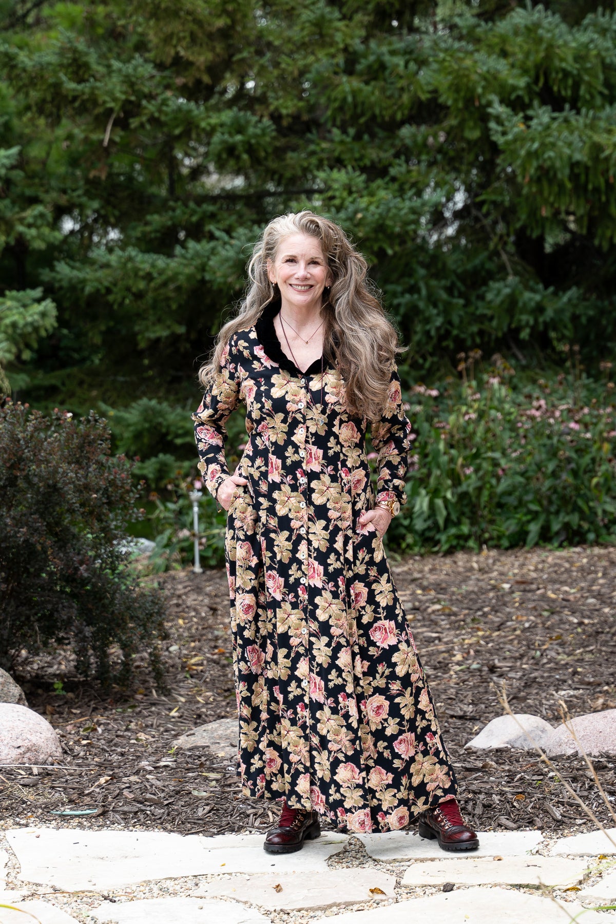 Melissa with long wavy blonde hair stands outdoors in a garden wearing a long-sleeve black maxi dress with a pink and beige floral pattern, paired with dark brown boots.