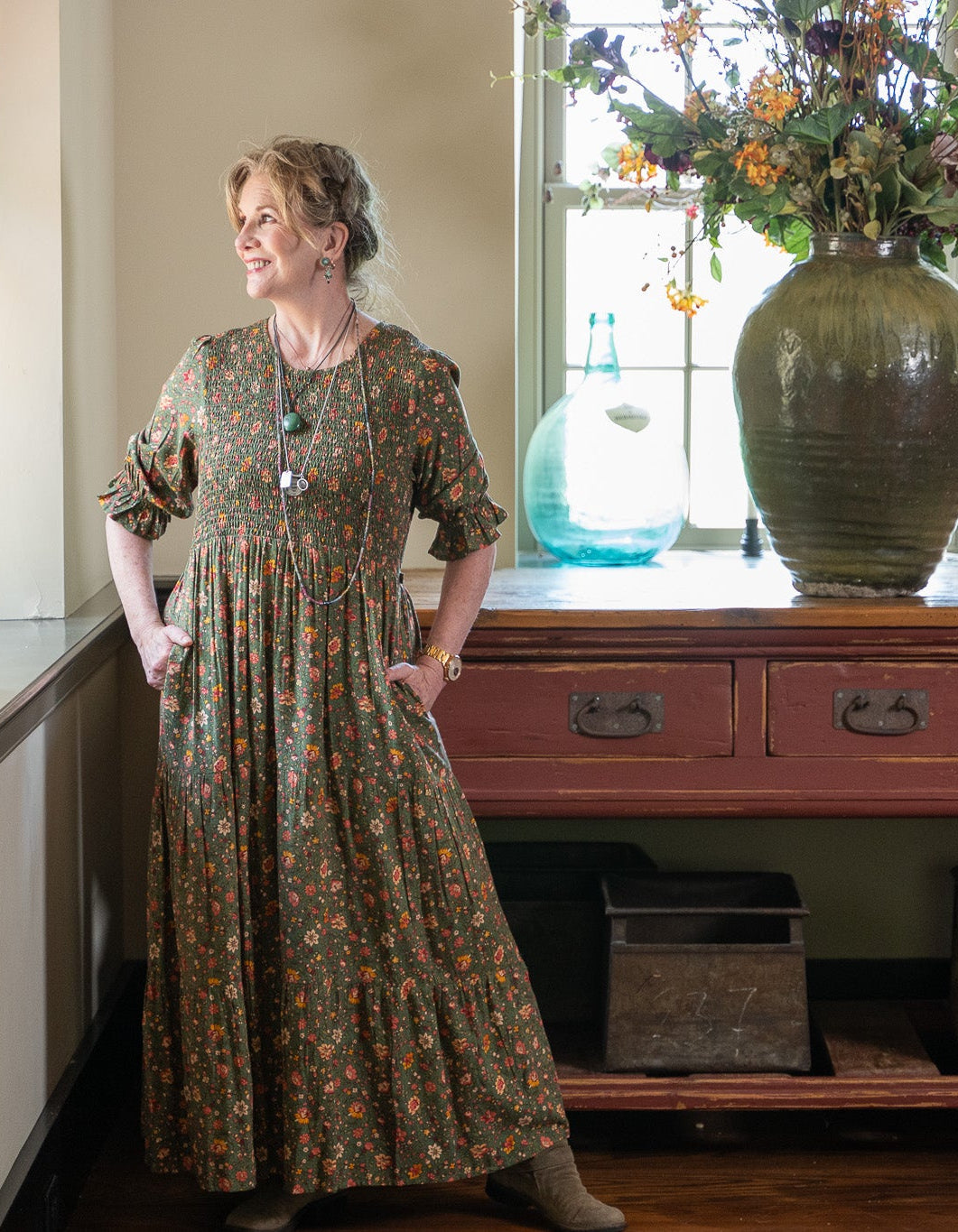 Melissa wearing a long-sleeved, knee-length floral print dress stands in a rustic indoor setting next to a window and a red wooden console table with a large green vase.