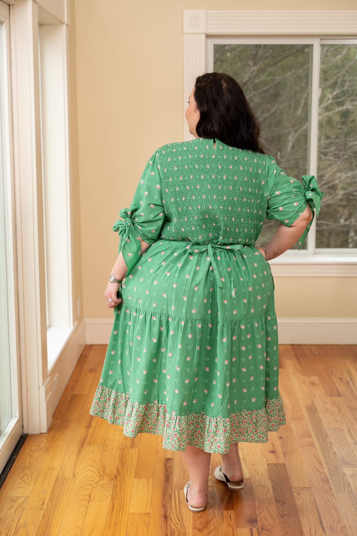 Woman wearing a green dress with floral pattern in a room with wooden floor and window.