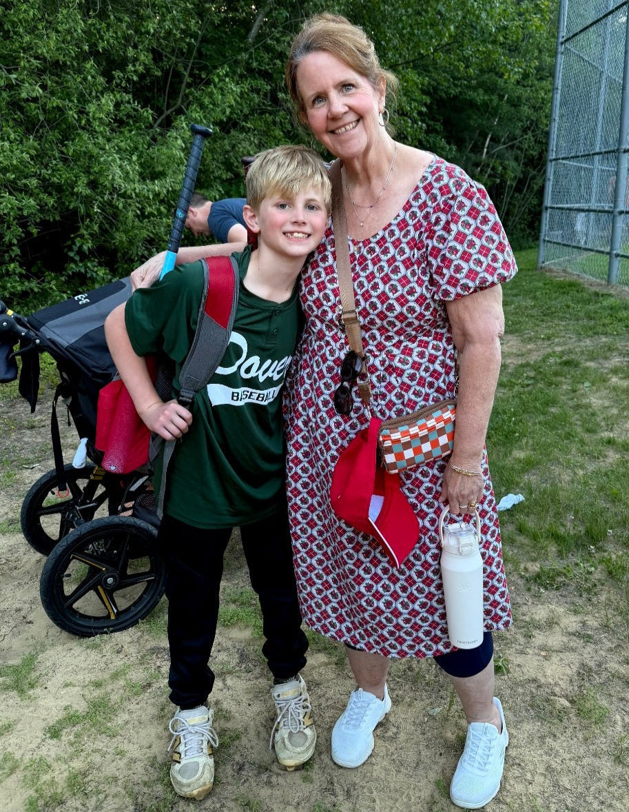 A woman and a young boy standing outdoors in front of a chain-link fence and trees. The woman wears a red, white, and blue patterned dress and white sneakers, holding a red bag and a white bottle.