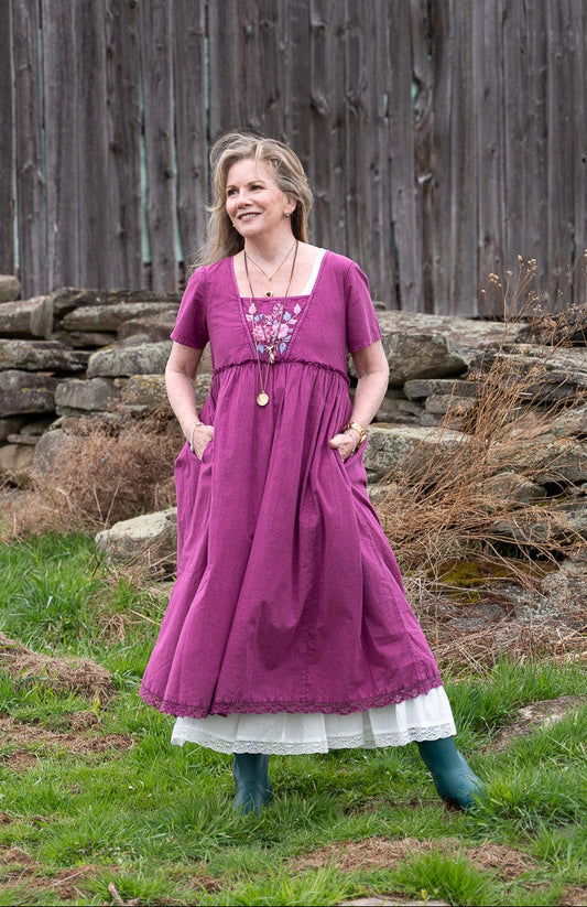 Melissa wearing a purple medieval-style linen dress stands in a grassy field in front of a weathered gray wooden barn, with a white dog on the left.
