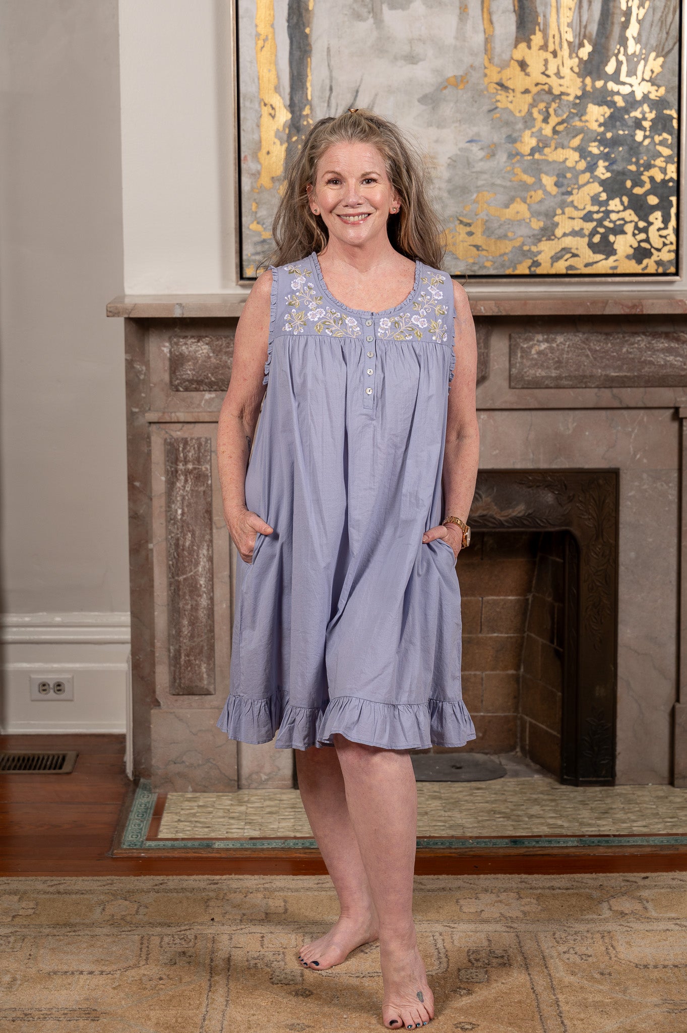 Melissa in a light blue dress standing in front of a fireplace with a decorative painting above it.
