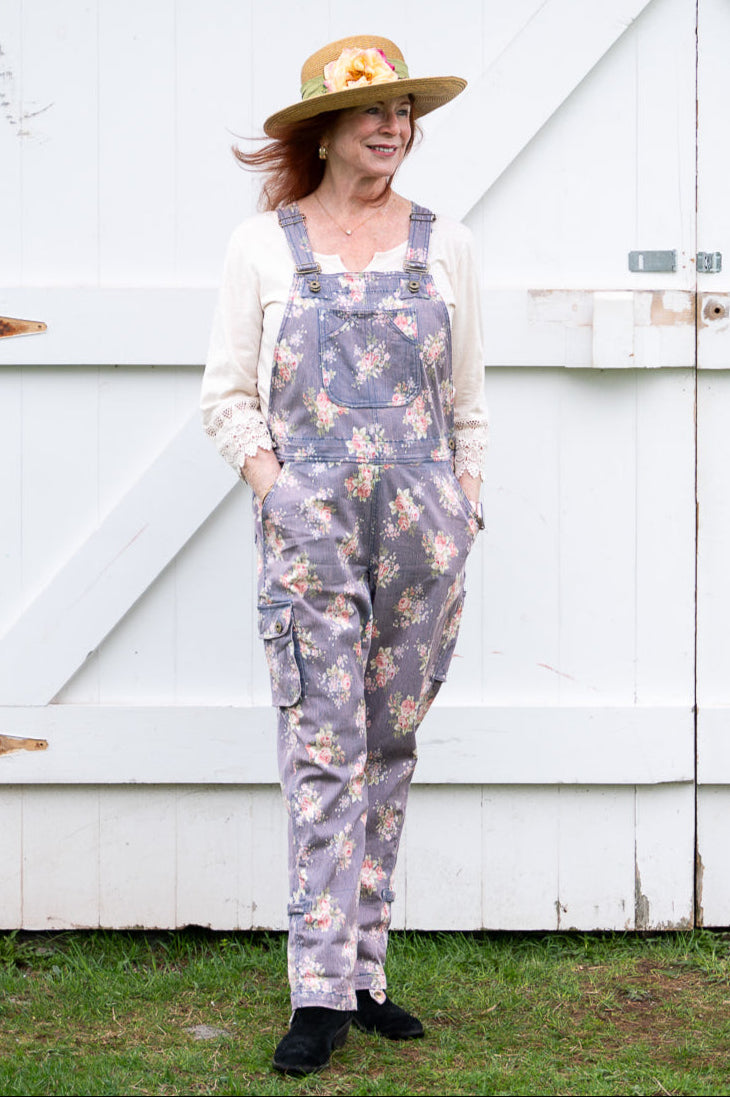 Sandy wearing floral overalls and a hat standing in front of a white wooden door.