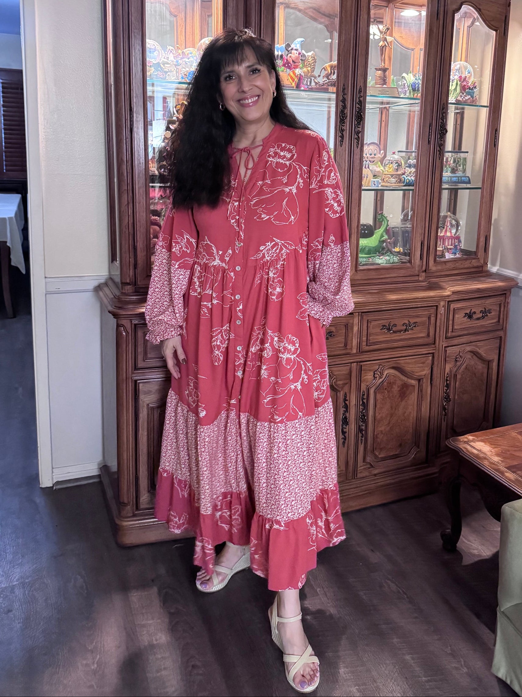A woman wearing a pink and white patterned maxi dress stands in a home interior next to a large dark wood hutch cabinet.
